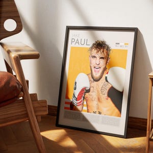 Puede incluir: Un póster enmarcado con un retrato de Jake Paul, un boxeador profesional, que lleva guantes de boxeo. El póster tiene un fondo amarillo con el texto "JAKE PAUL" en letras negras. El póster está enmarcado en negro.