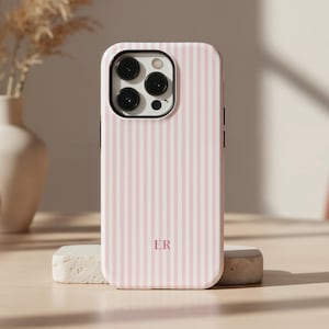 May include: A pink and white striped phone case with the initials "ER" in a serif font. The case is standing upright on a small, rectangular stone. The background includes a vase with dried flowers and a small, round vase.