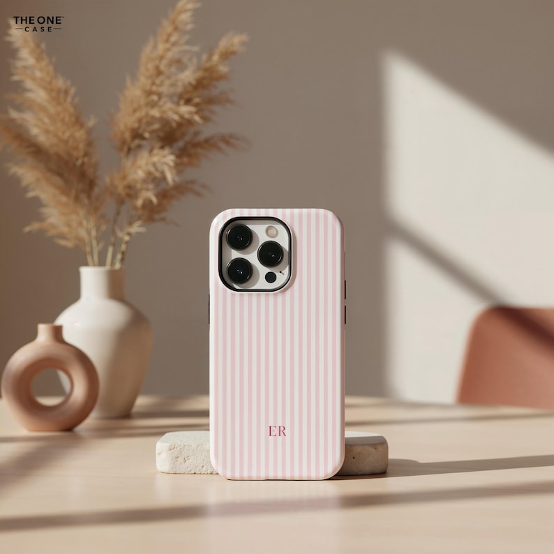 Pu&ograve; includere: Una custodia per telefono a righe rosa e bianche con le iniziali "ER" in un carattere serif. La custodia &egrave; in piedi su una piccola pietra rettangolare. Lo sfondo include un vaso con fiori secchi e un piccolo vaso rotondo.