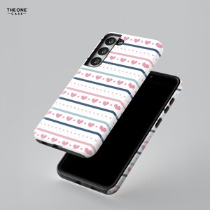 May include: A white phone case with a pink and blue striped pattern and pink hearts. The case is designed for a phone with a triple camera lens.