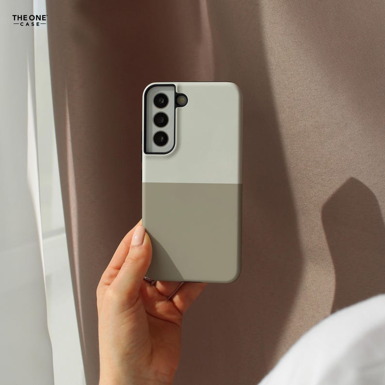 May include: A two-tone phone case in a vertical split design, held in a hand. The top half is a light beige, and the bottom half is a muted olive green. The phone case has a camera cutout with three lenses. The text "THE ONE -CASE-" is in the upper left corner.