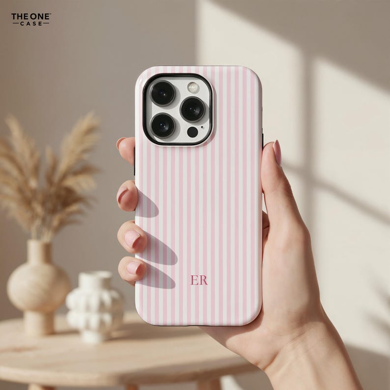Pu&ograve; includere: Una custodia per telefono a righe rosa e bianche con le iniziali "ER" in basso al centro. La custodia del telefono ha una protezione per l'obiettivo della fotocamera nera. Il marchio "THE ONE CASE" si trova nell'angolo in alto a sinistra.