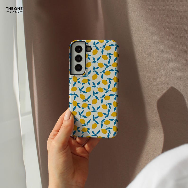 Op de afbeelding: Een witte telefoonhoes met een patroon van gele citroenen en blauwe bladeren. De telefoonhoes wordt in een hand gehouden. De tekst "THE ONE -CASE-" staat in de linkerbovenhoek.