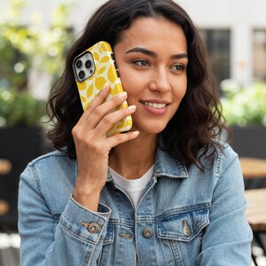 Op de afbeelding: Gele telefoonhoesje met een wit bladerpatroon. De telefoon wordt tegen het oor gehouden. De telefoonhoes heeft een zwarte cameralens. De tekst "THE ONE -CASE-" staat in de linkerbovenhoek.
