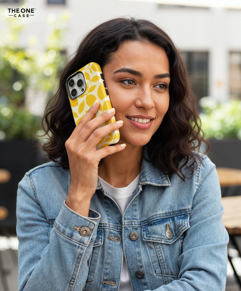 Op de afbeelding: Gele telefoonhoesje met een wit bladerpatroon. De telefoon wordt tegen het oor gehouden. De telefoonhoes heeft een zwarte cameralens. De tekst "THE ONE -CASE-" staat in de linkerbovenhoek.