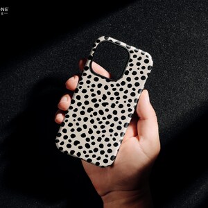 May include: A phone case with a cream-colored background and a pattern of black spots. The case has a cutout for the camera and is held in a hand. The text "THE ONE -CASE-" is in the upper left corner.