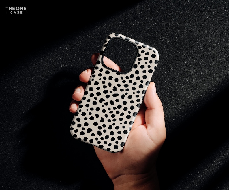 May include: A phone case with a cream-colored background and a pattern of black spots. The case has a cutout for the camera and is held in a hand. The text "THE ONE -CASE-" is in the upper left corner.