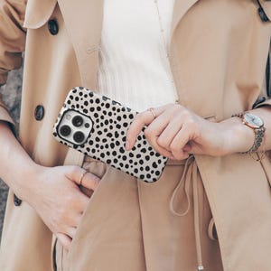 May include: A smartphone case with a black and white dalmatian spot pattern is held in front of a tan trench coat. The case has a camera cutout and is held by a person wearing a watch and rings. The coat has black buttons.