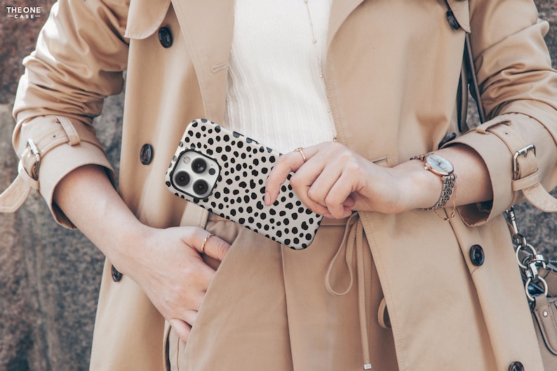 May include: A smartphone case with a black and white dalmatian spot pattern is held in front of a tan trench coat. The case has a camera cutout and is held by a person wearing a watch and rings. The coat has black buttons.