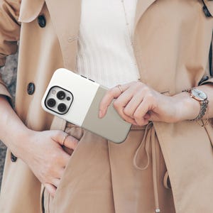 May include: A two-tone phone case in white and gray is held in front of a tan trench coat. The phone case has a camera module with multiple lenses. The coat has a belt, buttons, and a watch on the wrist.
