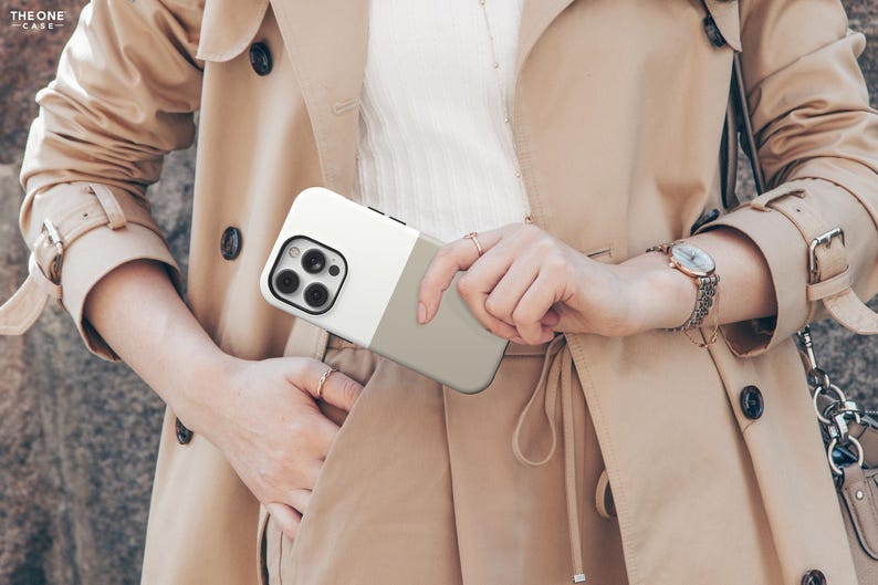May include: A two-tone phone case in white and gray is held in front of a tan trench coat. The phone case has a camera module with multiple lenses. The coat has a belt, buttons, and a watch on the wrist.