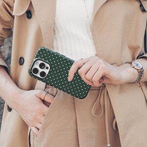May include: A green and white polka dot phone case with a camera lens cutout. The case is being held by a person wearing a beige trench coat.