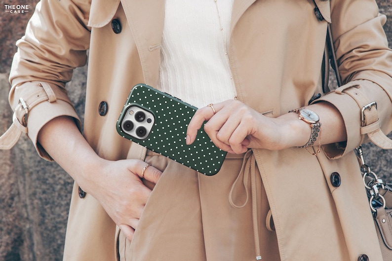 May include: A green and white polka dot phone case with a camera lens cutout. The case is being held by a person wearing a beige trench coat.