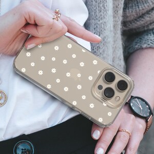 May include: A clear phone case with a white daisy pattern. The case is on a gold iPhone with a triple lens camera.