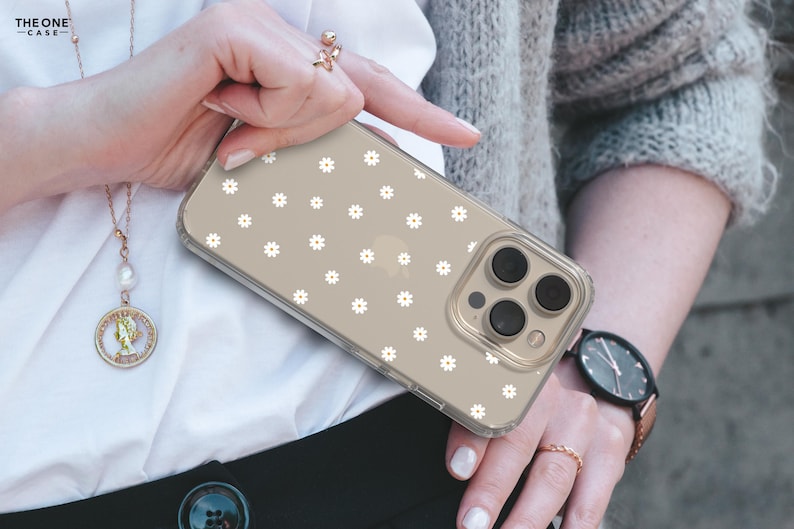 May include: A clear phone case with a white daisy pattern. The case is on a gold iPhone with a triple lens camera.