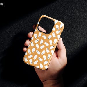 May include: A phone case with a repeating pattern of white ghost illustrations on a tan background. The case has a black border and the text "THE ONE -CASE-" in the upper left corner.