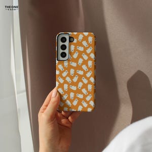 May include: A phone case with a repeating pattern of white cartoon ghosts on an orange background. The phone case is held in a hand. The phone case has a camera module with three lenses. The text "THE ONE -CASE-" is in the upper left corner.