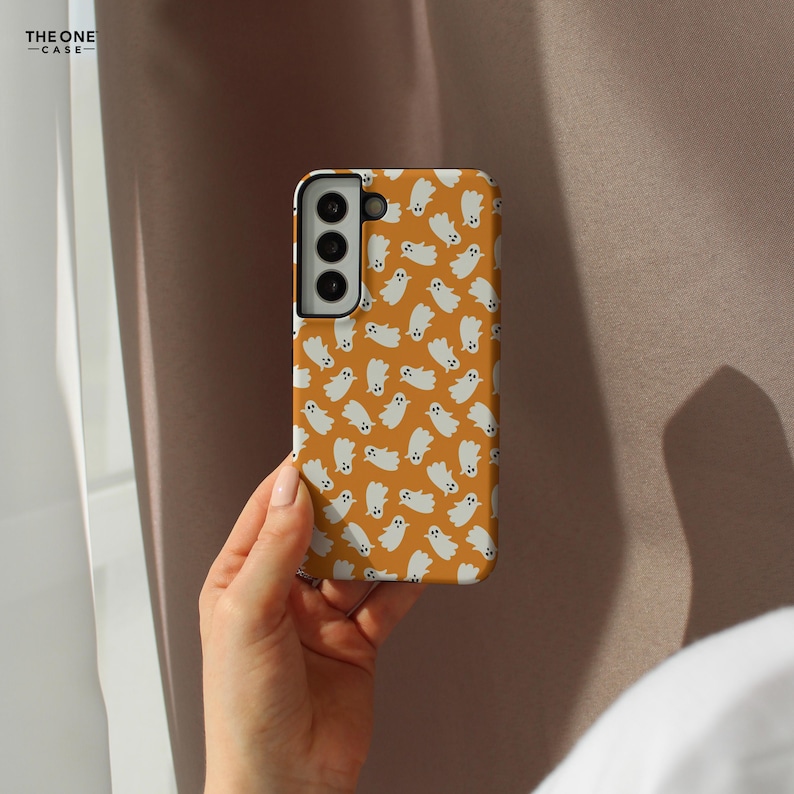 May include: A phone case with a repeating pattern of white cartoon ghosts on an orange background. The phone case is held in a hand. The phone case has a camera module with three lenses. The text "THE ONE -CASE-" is in the upper left corner.