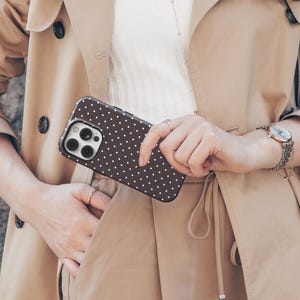 May include: A brown phone case with a white polka dot pattern. The phone case is held in front of a tan trench coat. The image also shows a watch with a silver band and a small purse.