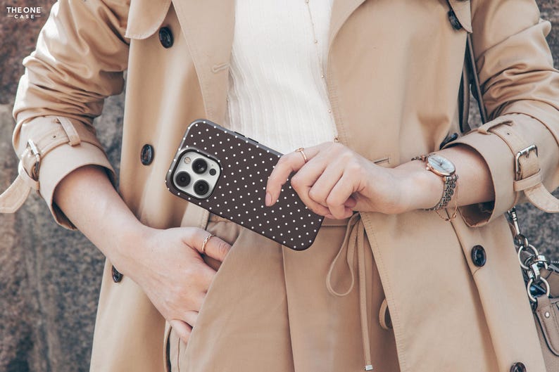 May include: A brown phone case with a white polka dot pattern. The phone case is held in front of a tan trench coat. The image also shows a watch with a silver band and a small purse.