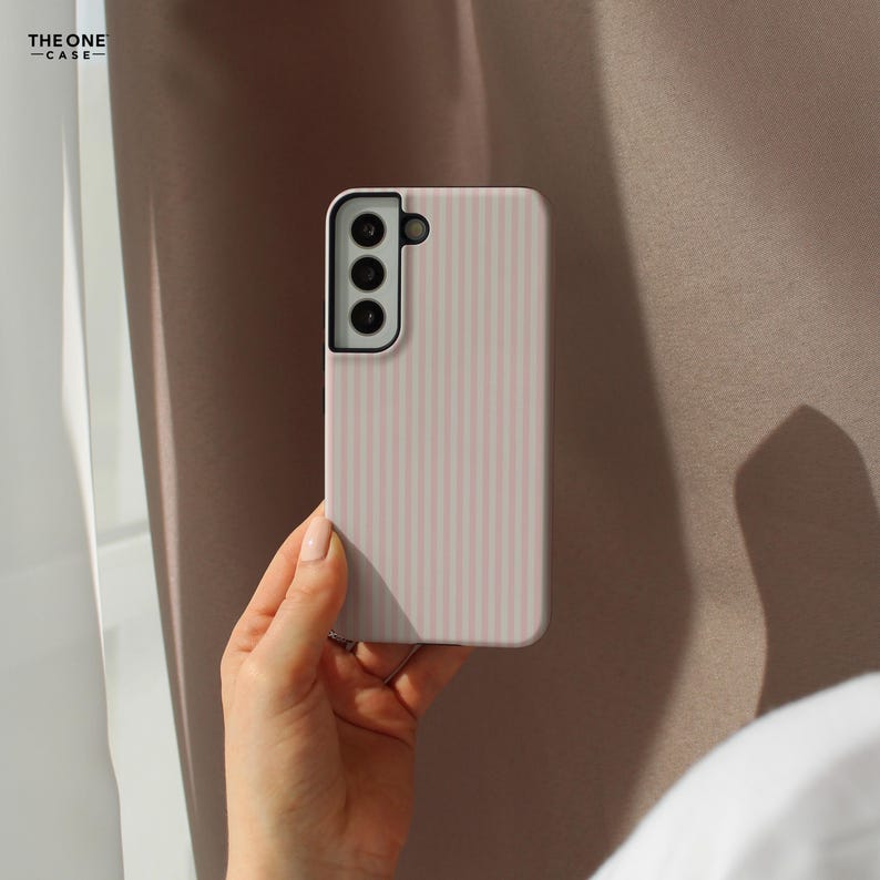 Pu&ograve; includere: Custodia per telefono rosa chiaro con strisce verticali e un ritaglio per la fotocamera. La custodia &egrave; tenuta in mano, con il marchio "THE ONE -CASE-" visibile in alto a sinistra. Lo sfondo &egrave; di colore neutro.