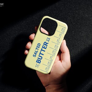 Pu&ograve; includere: Una custodia per telefono giallo chiaro con le parole "SALTED BUTTER" in blu, insieme a segni di misurazione. La custodia ha anche il testo "GRADE AA" e "NET WT 113g". La custodia del telefono &egrave; tenuta in una mano.