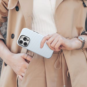 Pode incluir: Uma capa de telefone listrada em azul claro e branco, segurada por uma pessoa vestindo um trench coat bege. A capa do telefone tem um recorte para a c&acirc;mera e uma borda preta. A pessoa tamb&eacute;m est&aacute; usando um rel&oacute;gio com pulseira prateada.