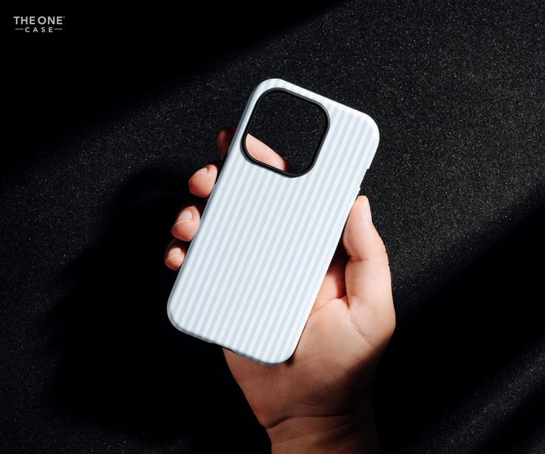 Pode incluir: Capa de telefone azul claro com um padr&atilde;o listrado, segurada na m&atilde;o. A capa tem um recorte preto para a c&acirc;mera e o texto "THE ONE -CASE-" no canto superior esquerdo. O fundo &eacute; escuro.