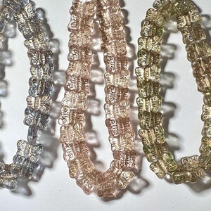 May include: Three strands of butterfly-shaped beads in shades of blue, pink, and green. The beads are translucent, allowing light to pass through, and are strung together to form necklaces or bracelets. The beads are likely made of glass or a similar material.