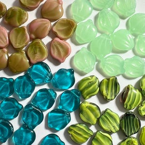 May include: A collection of colorful glass beads in various shapes. The beads are in shades of pink, green, blue, and yellow. Some beads have a shell-like shape, while others are more rounded. The beads are arranged on a white surface.