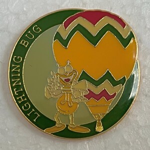 May include: A round metal enamel pin featuring a cartoon lightning bug character standing next to a large, colorful hot air balloon. The balloon has yellow, green, and pink chevron patterns. The text LIGHTNING BUG is printed along the green border.