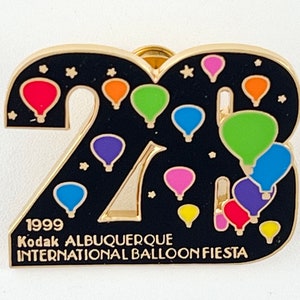 May include: A gold-toned pin featuring the number "23" with a black background and colorful hot air balloons. The pin includes the text "1999 Kodak Albuquerque International Balloon Fiesta".