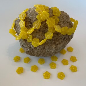 May include: A string of yellow flower-shaped glass beads wrapped around a rough, gray rock.  More yellow flower beads are scattered on a white surface.