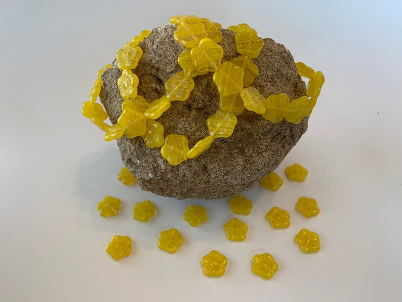 May include: A string of yellow flower-shaped glass beads wrapped around a rough, gray rock.  More yellow flower beads are scattered on a white surface.