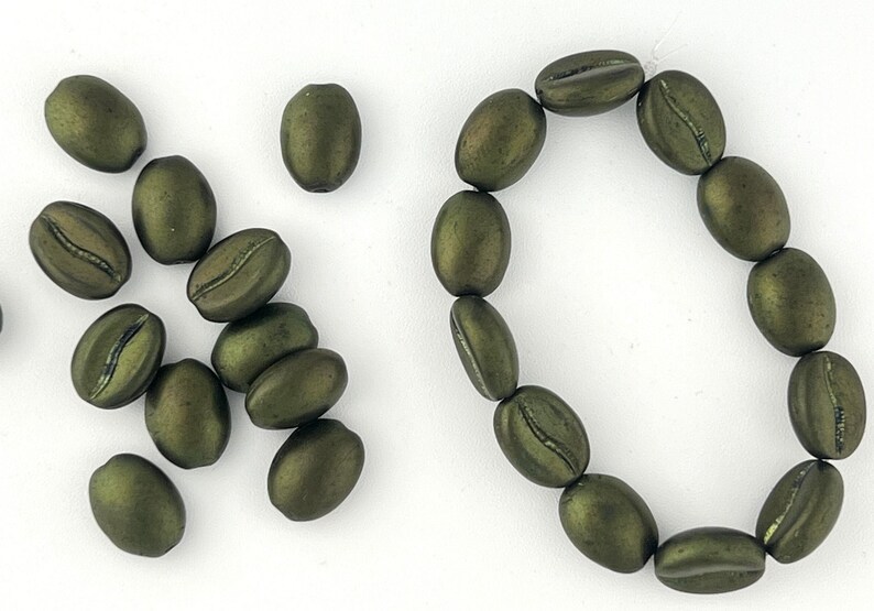 May include: A collection of green coffee bean shaped beads. Some beads are arranged in a circle, while others are scattered on a white surface.