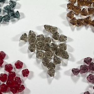 May include: A collection of small, translucent, flower-shaped glass beads arranged in separate color groups. The clusters include shades of smoky grey, deep red, amber, dark blue, and purple, each featuring a central hole for threading.