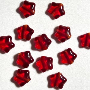 May include: A collection of eleven translucent, star-shaped beads. Each bead is a vibrant red with a subtle internal stripe and a glossy finish. The beads are scattered against a white background, showcasing their shape and color.