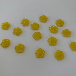May include: A collection of 15 yellow flower-shaped buttons. The buttons are made of a shiny, translucent material and have a slightly raised design.