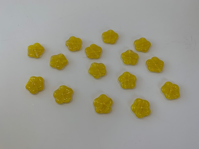May include: A collection of 15 yellow flower-shaped buttons. The buttons are made of a shiny, translucent material and have a slightly raised design.