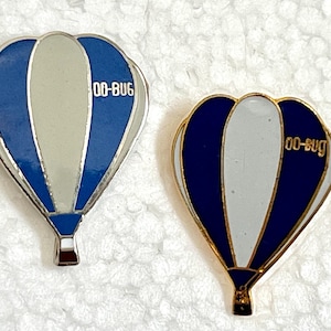 May include: Two enamel pins shaped like hot air balloons. The left pin features blue and light gray panels with the text 00-BUG in white. The right pin has dark blue and white panels with the text 00-bug in gold. Both pins have metallic outlines.
