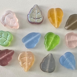 May include: A collection of twelve small, leaf-shaped glass beads in various translucent and frosted colors, including shades of pink, green, blue, yellow, and gray. Each bead features a molded vein pattern on the surface, creating a textured appearance.
