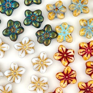May include: Assortment of flower-shaped glass beads in various colors, including white, blue, red, and green. The beads have a gold-colored center and are arranged on a white surface.
