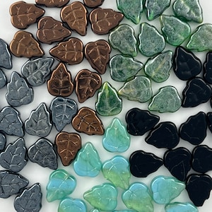 May include: Assortment of leaf-shaped beads in various colors, including bronze, gray, green, black, and teal. The beads have a detailed vein pattern and are scattered across a white surface. These beads are suitable for jewelry making and craft projects.
