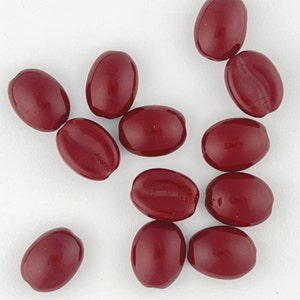 May include: A collection of 11 smooth, oval-shaped beads in a deep burgundy red color. The beads are scattered on a white surface.