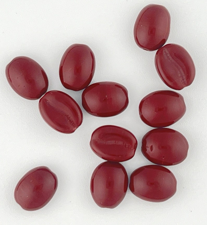 May include: A collection of 11 smooth, oval-shaped beads in a deep burgundy red color. The beads are scattered on a white surface.