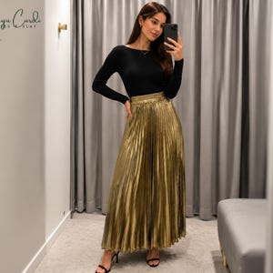May include: A woman wearing a black long-sleeve top and a gold pleated skirt. The skirt is floor-length and has a metallic sheen. The image is taken indoors, with a neutral background.