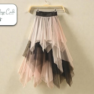 May include: A layered tulle skirt in black, pink, and beige hangs from a wooden hanger. The skirt has an elasticated waistband and an asymmetrical hemline. The brand logo is in the upper left corner.