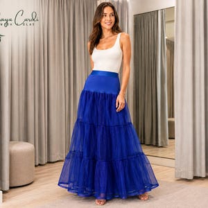 May include: A woman wearing a white tank top and a long, tiered, royal blue tulle skirt. The skirt has a wide, solid blue waistband. The brand logo "Elayu Curdi" is visible in the upper left corner. The background includes neutral-colored curtains and a mirror.