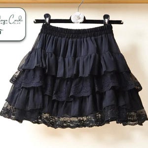 May include: Black ruffled skirt with lace trim, hanging on a black hanger. The skirt has multiple layers of fabric, creating a voluminous silhouette. The elastic waistband is visible. The brand logo is in the upper left corner.