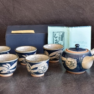 Può includere: Un set da tè in ceramica composto da una teiera con manico laterale e quattro tazze abbinate, decorate con motivi botanici blu scuro e crema e una finitura smaltata crepata. Una scatola blu scuro e un foglietto illustrativo con testo giapponese.